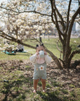 A toddler in a floral romper stands barefoot on grass, sipping from a Mushie Trainer Sippy Cup with easy-grip handles under a blooming tree with white flowers on a sunny day in the park.