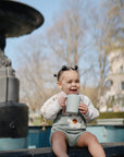 A smiling baby sits by a fountain, holding a Mushie Trainer Sippy Cup with easy-grip handles. Dressed in a flower-patterned sweater and overalls, the baby is framed by trees and a building in the background.