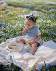 A young child with small buns in their hair sits on a blanket in a grassy field, eating fruit from a divided plate and wearing light-colored clothes plus the mushie Muslin Bib, enjoying a sunny day among purple flowers.