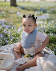 A young child with braided hair sits on a blanket among purple flowers, holding a snack. Nearby are a divided plate and the FRIGG Butterfly Anatomical Silicone Pacifier 2-Pack (6-18 Months), adding to the playful scene.