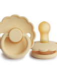 Two FRIGG Daisy Natural Rubber Pacifiers in Pale Daffodil are shown: one flat with the nipple up, the other upright with the handle forward. Both are limited edition and display scalloped edges.