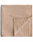 The mushie Organic Cotton Muslin Swaddle Blanket in Pale Taupe is shown folded with a visible grid texture and one corner turned over, displayed on a white background.
