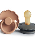 The FRIGG Daisy Natural Rubber Pacifier 2-Pack includes two daisy-shaped pacifiers, one light brown and one dark gray, each with a round handle and natural rubber nipple, branded FRIGG at the base.