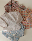 Four mushie Long Sleeve Bibs—pink abstract, colorful squares, cherry print, and blue floral—are displayed overlapping on a light surface. Each bib is water resistant, has a wide pocket, and is machine washable for everyday convenience.