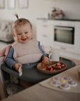 A smiling toddler with a ponytail sits in a high chair using the mushie Stage 2 Cambridge Blue Feeding Kit, which includes baby utensils, a training cup with straw, and a plate of snacks on her tray in a modern kitchen.