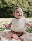A smiling baby in a light brown outfit sits outdoors on a blanket, wearing the mushie Silicone Baby Bib with an adjustable neck strap, surrounded by toys and blurred green trees in the background.