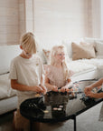 Three blonde-haired children build with the mushie HEX Connect Building Toy Set—a fun STEM activity for spatial skills—on a black coffee table in a bright, cozy living room with a cream sofa and wooden walls.