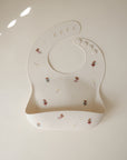 The mushie Silicone Baby Bib in cream, featuring an adjustable neck strap and bottom pocket, is decorated with fairy and star illustrations and displayed flat on a light background.