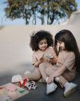 Two young children sit outside on concrete, smiling and wearing matching beige outfits, while playing with mushie Silicone Animal Finger Puppets that help develop fine motor skills. Trees can be seen in the background.