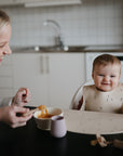 A smiling baby in a high chair is fed by an adult woman using the mushie Stage 1 Bees Feeding Kit. A bowl of food and cup—mealtime essentials from the kit—are on the table beside them.