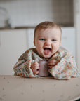 A smiling baby in a modern kitchen wears the mushie Long Sleeve Bib with a floral pattern, holding a small cup while seated in a high chair.