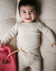 A smiling baby in a mushie Two Piece Set lies on a cushioned surface, holding a beaded pacifier clip. The cozy long sleeve shirt and pants ensure comfort, with a pink blanket partially visible beside the baby.