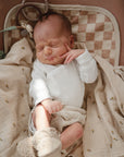 A newborn in a white outfit and knitted booties rests on a Mushie Changing Pad Liner beneath a light beige blanket, eyes closed, with one hand gently touching their cheek.