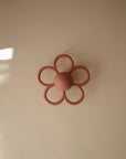 The mushie Daisy Rattle Teether features a mauve pink, flower-shaped design with a round center and five looped petals, displayed on a beige surface under soft lighting with a shadow in the corner.