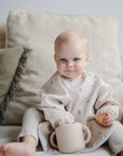 A baby with light hair sits on a beige couch, wearing a light-colored outfit. The baby holds a cup and cookie from the mushie Deluxe Vanilla Confetti/Cambridge Blue Feeding Kit, looking at the camera with a slight smile.