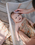A baby wrapped in a floral towel smiles up from a clear tub as an adult gently brushes their hair with mushie Muslin Cotton Washcloths. The scene features a wooden floor and a patterned cloth nearby.
