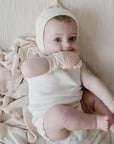 A baby in a white outfit and knit bonnet lies on a beige blanket, happily chewing the mushie Rainbow Teething Mitten—a soothing silicone glove designed for teething comfort.