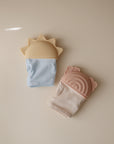 Two mushie Rainbow Teething Mittens rest on a beige surface: one light blue with a sun-shaped yellow top, and one off-white with pink, bear-ear accents—both designed to soothe babies’ gums.