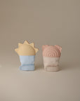 Two Rainbow Teething Mittens by mushie stand upright on a neutral background—one light blue with a sun-shaped top, the other beige with a pink top featuring rainbow and ear-like shapes. These silicone gloves gently soothe babies’ gums.