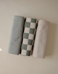 Three Mushie Changing Pad Liners—one solid light gray, one green and white checkered, and one solid cream—made from organic cotton muslin, are neatly rolled side by side on a light background.