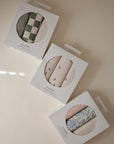 Three boxes of Mushie Changing Pad Liners are arranged on a light surface, each showing a unique pattern: green-checkered, beige with small flowers, and blue floral on pale fabric.