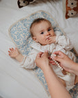 A baby in white lies on Mushie Changing Pad Liners with a blue and white pattern, sucking their fingers while an adult holds a beaded pacifier clip nearby.