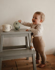 A smiling baby wearing a long-sleeve shirt and brown pants reaches for a tipped-over mug and the mushie Bluey x Mushie Snack Cup atop a gray step stool, exploring these feeding essentials in a cozy home setting.