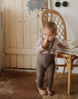 A toddler stands barefoot on a rug, eating an apple from the mushie Bluey x Mushie Snack Cup. They lean against a wicker chair with the cup on it, in front of a rustic wooden door—a cozy scene for exploring baby feeding essentials.