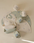 Two mushie Bluey x Mushie Silicone Baby Bibs, three sippy cups, and two placemats with pastel star and animal prints are arranged on a light surface in soft green, white, and pale pink shades for a charming nursery look.