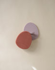 A mauve mushie wall hook with a curved, minimalist design holds a red Cradle Cap Brush with soft silicone bristles, set against a neutral beige background—an essential for gentle baby bath routines.