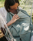 A woman sits outdoors in a hammock, holding the mushie Muslin Nursing Cover close to her chest as sunlight filters through the trees around her.