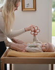 A woman stands beside a changing table, holding the mushie Daisy Rattle Teether above a baby in knitted clothes and a hat, who is lying on a mustard-colored blanket and reaching for the toy.