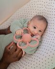 A baby in a green knit outfit lies on a soft dotted blanket, holding the mushie Daisy Rattle Teether, with an adult hand gently resting nearby.