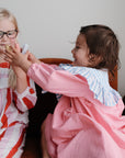 Two young children sit together; one smiles in glasses and a red striped outfit while the other, in a pink dress, holds up mushie Silicone Animal Finger Puppets—fun bath toys that encourage fine motor skill development.