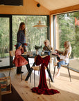 A woman and three children enjoy a meal with mushie Square Dinnerware Plates in a sunlit room with large windows. A chair decorated with a large red bow sits in the foreground, and trees can be seen outside.
