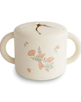 The mushie Snack Cup is a cream-colored toddler snack container with two handles and a spill-proof lid featuring petal-like openings for easy snack access; it’s decorated with pink flowers.