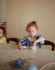 Two young children with curly hair sit at a table. One, in a blue and white sweater, plays with mushie Silicone Baby Food Containers, while the other, in a yellow sweater, rests their elbow on the table and looks away.