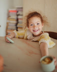 A smiling young child in a yellow and white sweater sits at a table, holding a mushie Silicone Baby Food Container. Stacks of books are visible in the background.