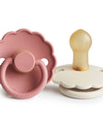 The FRIGG Daisy Natural Rubber Pacifier 2-Pack includes two scalloped pacifiers: one pink with a round handle (front view) and one cream featuring a natural rubber latex nipple (side view).