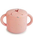 The mushie Snack Cup in Powder Pink Confetti is a silicone toddler snack container with two handles and a flexible slit lid, perfect for mess-free, spill-proof snacking on the go.