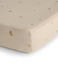 Close-up of the mushie Mini Muslin Crib Sheet, made from 100% cotton in beige with small brown rainbow and dot patterns, shown covering a crib mattress corner on a white background.