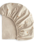 The mushie Stretchy Crib Sheet is shown partially folded on a white background. It’s beige with elastic edges and features a pattern of small, repeating brown rainbows.