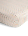 A close-up of the mushie Extra Soft Muslin Crib Sheet: breathable, with pastel orange, yellow, green, and blue vertical stripes on white, neatly fitted over a mattress corner.