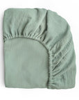 The mushie Extra Soft Muslin Crib Sheet is a light green, textured fitted sheet with elastic edges, neatly folded on a white background—ideal as breathable baby bedding or a soft muslin crib sheet.
