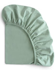 The mushie Stretchy Crib Sheet, crafted from soft organic cotton in a gentle green shade, is partially folded with elastic edges and a smooth, breathable surface displayed against a white background.