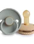 The Mushie FRIGG Rope Natural Rubber Pacifier 2-Pack features a soft grayish-blue and a beige pacifier, each with rope-like edges and ring handles—baby essentials crafted from smooth natural rubber latex.
