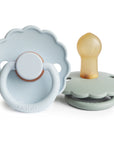 The FRIGG Daisy Natural Rubber Pacifier 2-Pack features two light blue, daisy-shaped pacifiers with scalloped edges and the FRIGG brand name embossed on the front. One displays the natural rubber nipple, while the other faces forward.