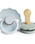 The FRIGG Daisy Natural Rubber Pacifier 2-Pack features two pastel pacifiers with scalloped, daisy-shaped edges—one front-facing with a round handle, the other displaying its natural rubber nipple. Both are branded “FRIGG.”.