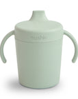 The Mushie Trainer Sippy Cup in light green features easy-grip handles, a spout lid, and the embossed Mushie logo on the front. This non-toxic sippy cup is perfect for little hands.