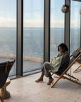 A person sits barefoot in a reclining chair by large windows, wrapped in a green mushie Muslin Nursing Cover, gazing out at the water with flags and a distant shoreline visible outside.